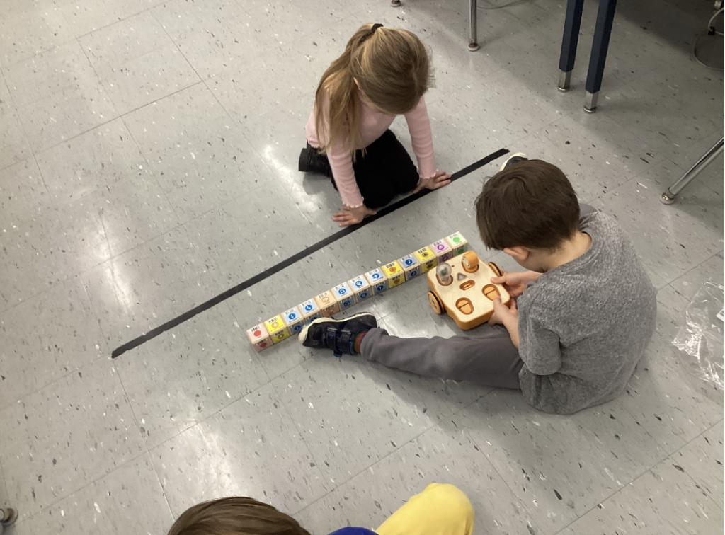 Two students sit on a classroom floor, working with a KIBO robot and its coding blocks. 